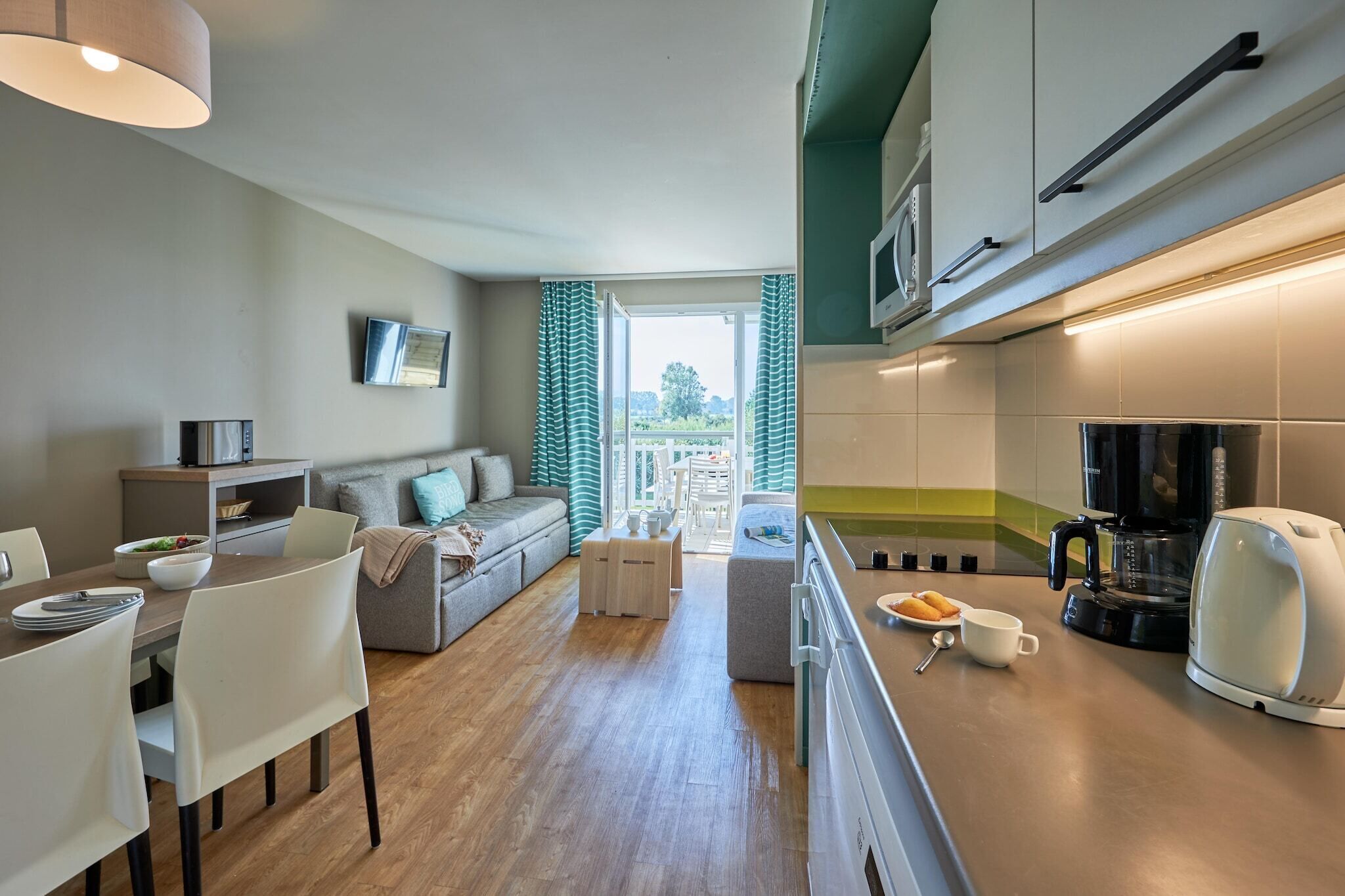 A modern kitchen with a microwave, coffee maker, and kettle. There is a dining area with a table and chairs, and a living area with a sofa and a television.
