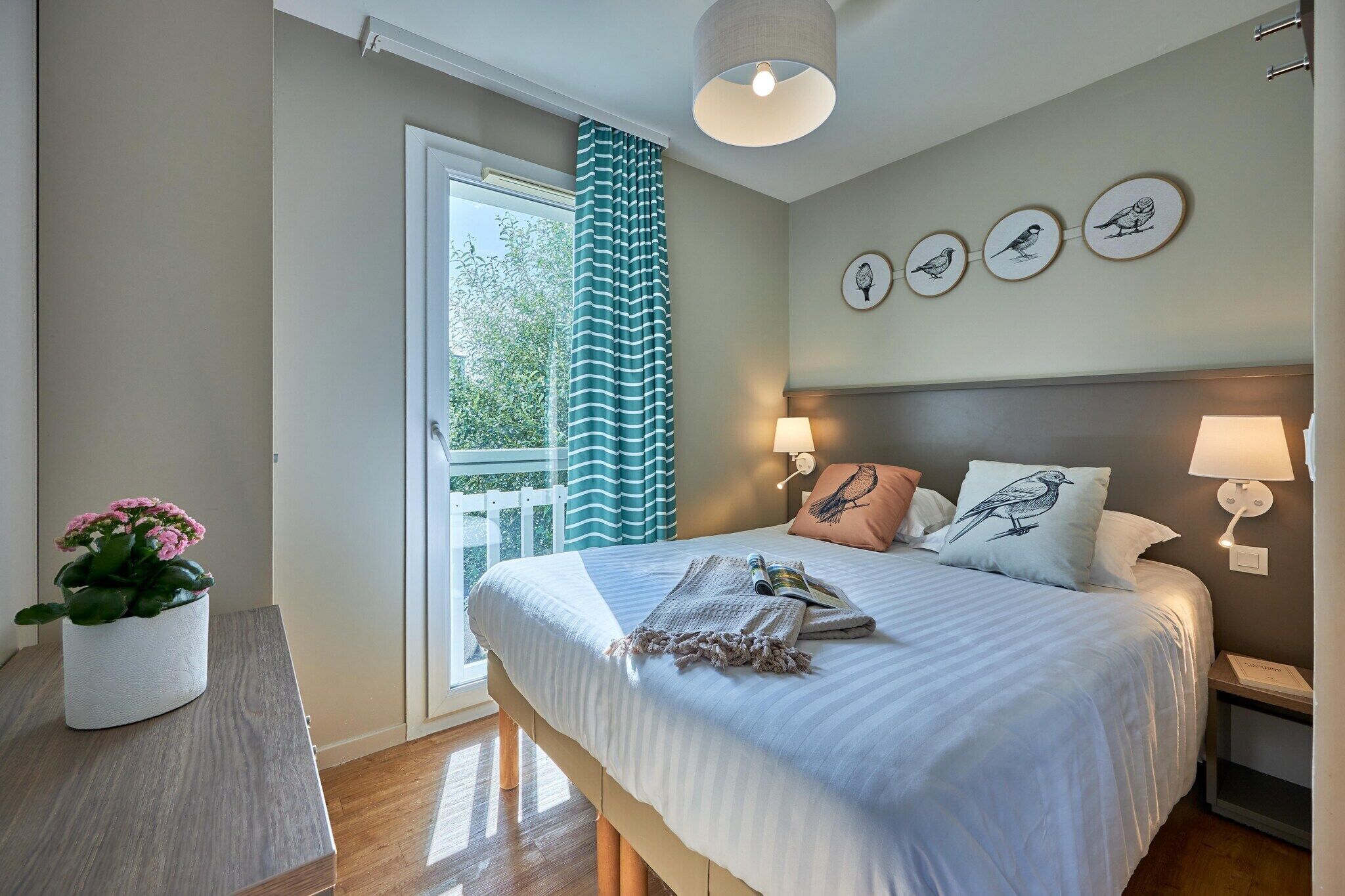 Une chambre à coucher avec un lit, des lampes de chevet, une table de chevet, une fenêtre avec des rideaux, une plante en pot et des œuvres d’art mural.