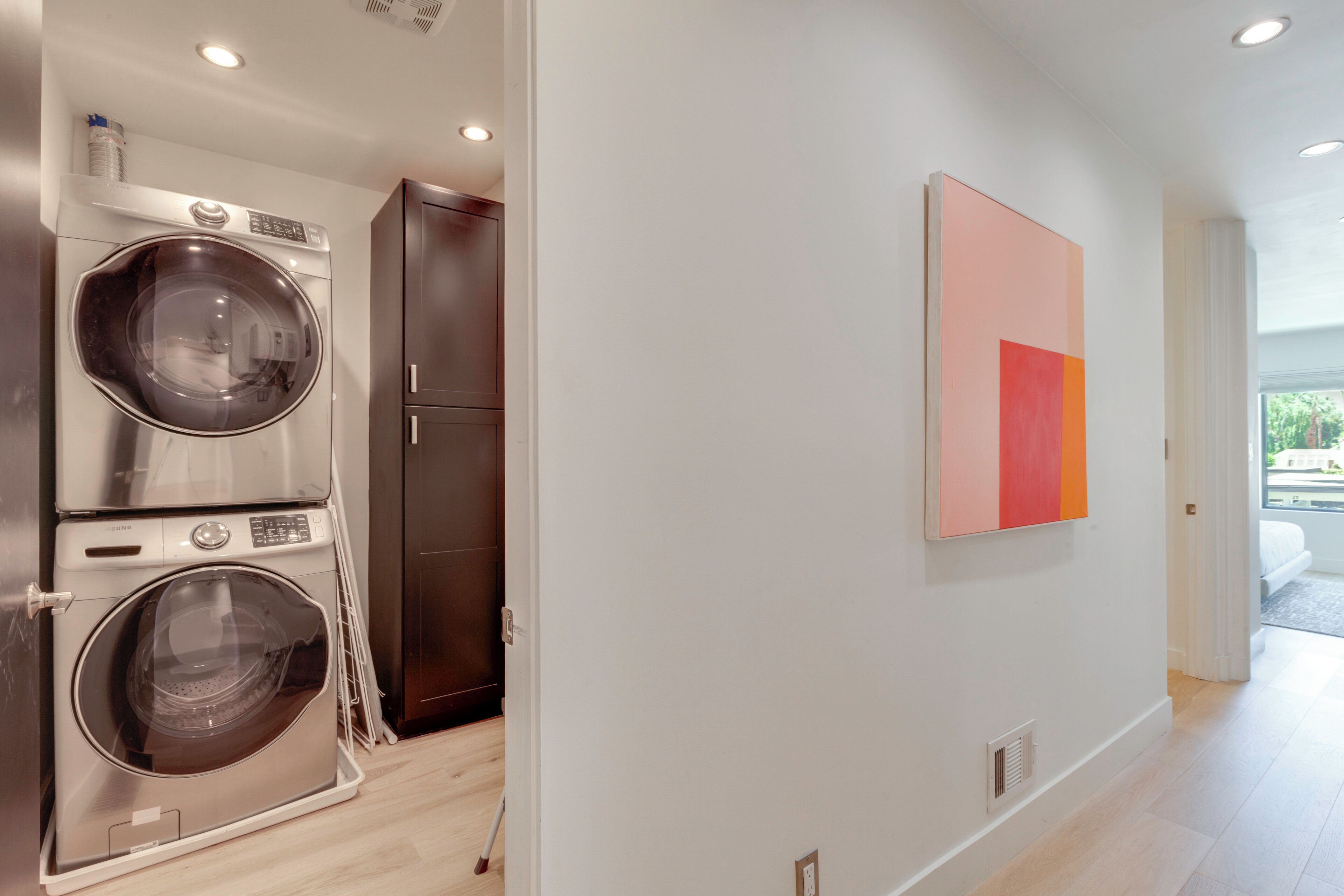 Une buanderie moderne avec des lave-linge et des sèche-linge superposés, un meuble en bois foncé et un mur épuré orné d’une seule œuvre d’art abstraite.