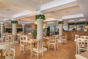 Un vaste espace dédié à la salle à manger, avec de nombreuses tables et chaises, un bar doté d’étagères remplies de bouteilles, et des plantes décoratives disposées sur des piliers.
