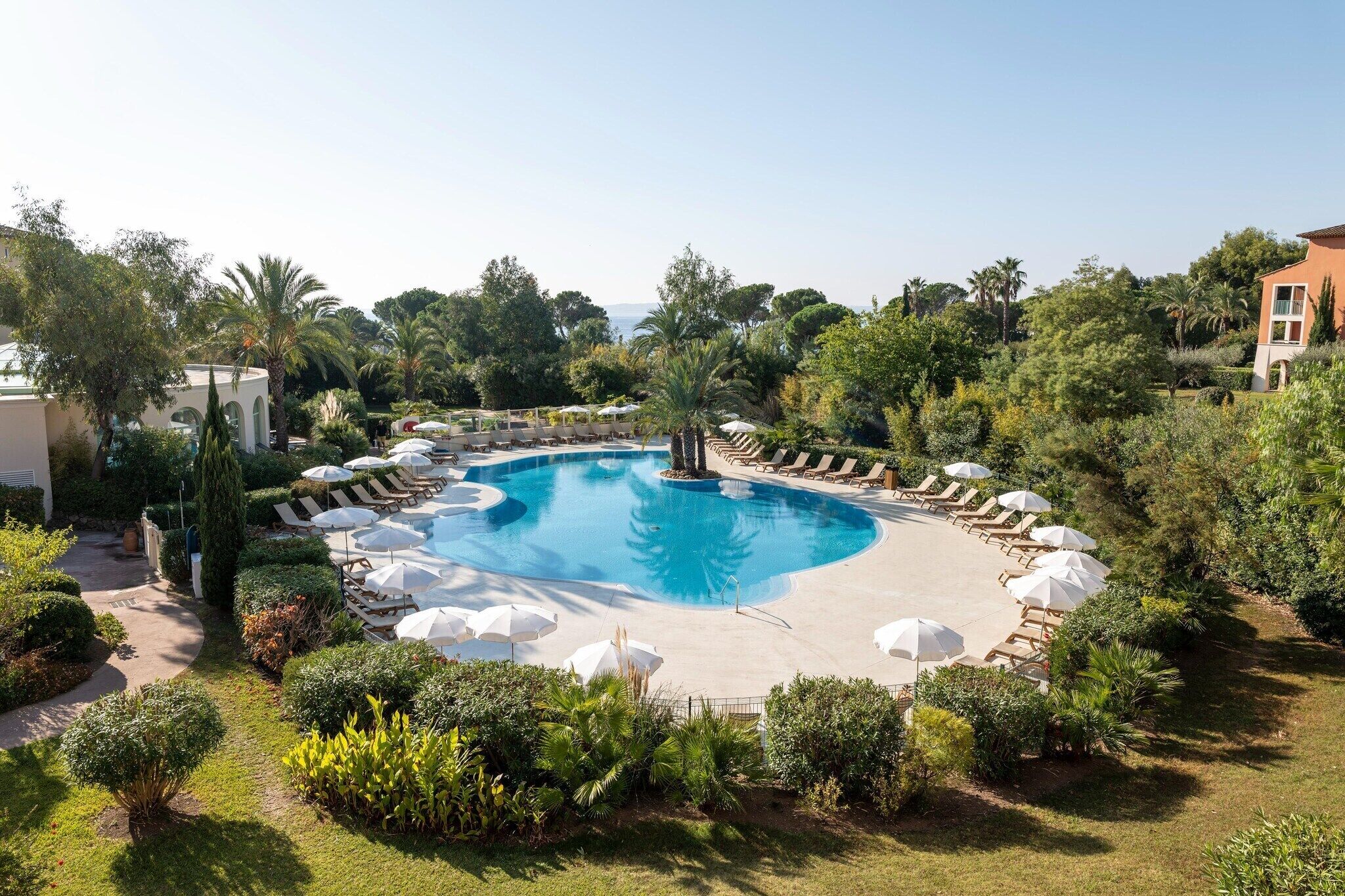 Ein kreisförmiger Pool mit Liegestühlen und Sonnenschirmen, umgeben von Grünflächen und einem Gebäude.