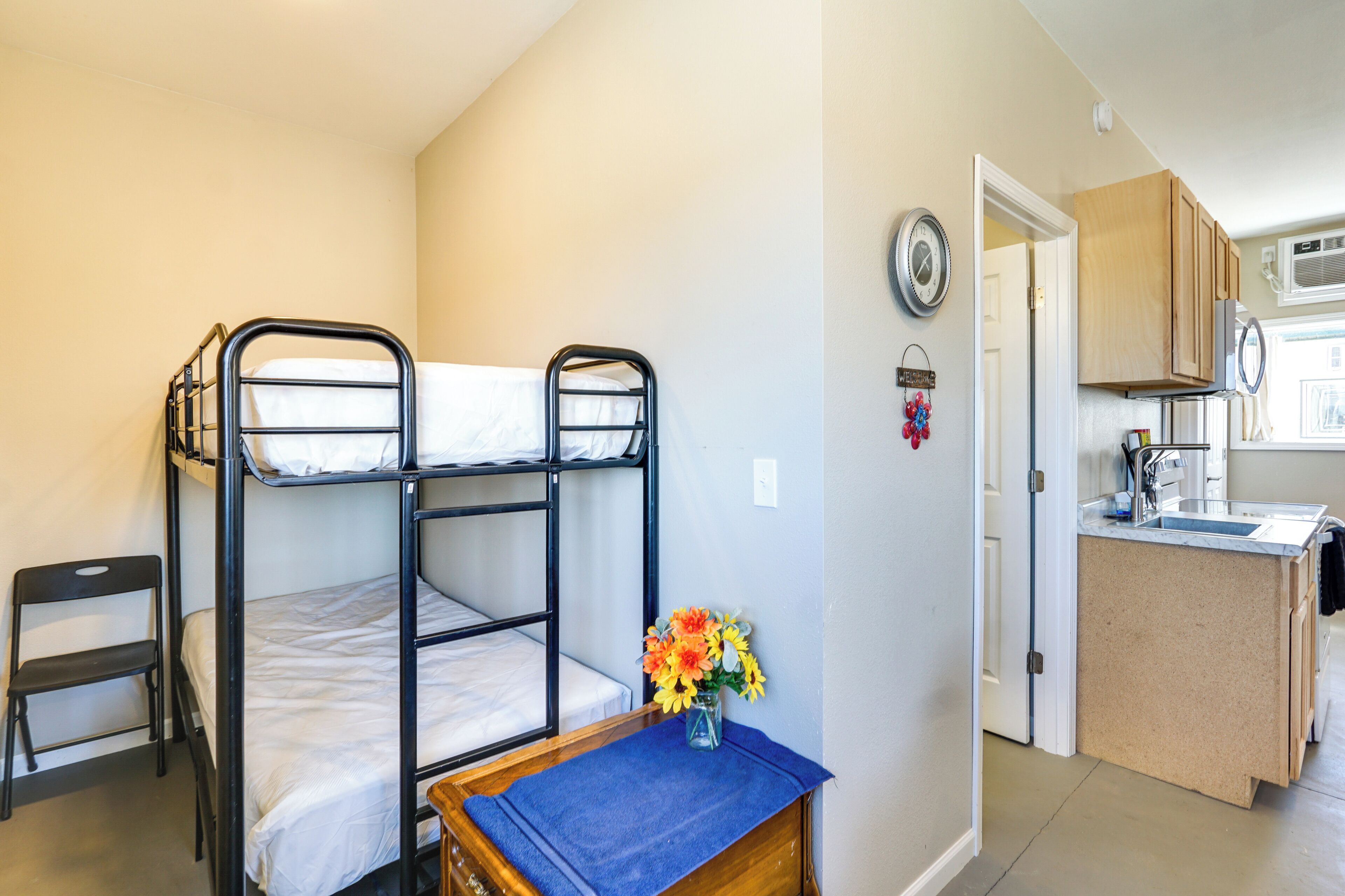 A bunk bed, a small table with a vase, a chair, and a kitchen area with cabinets and a sink.