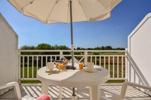 Un tavolo bianco con cibi per la colazione su un balcone dotato di ringhiera bianca, cielo azzurro limpido e area verde sullo sfondo.