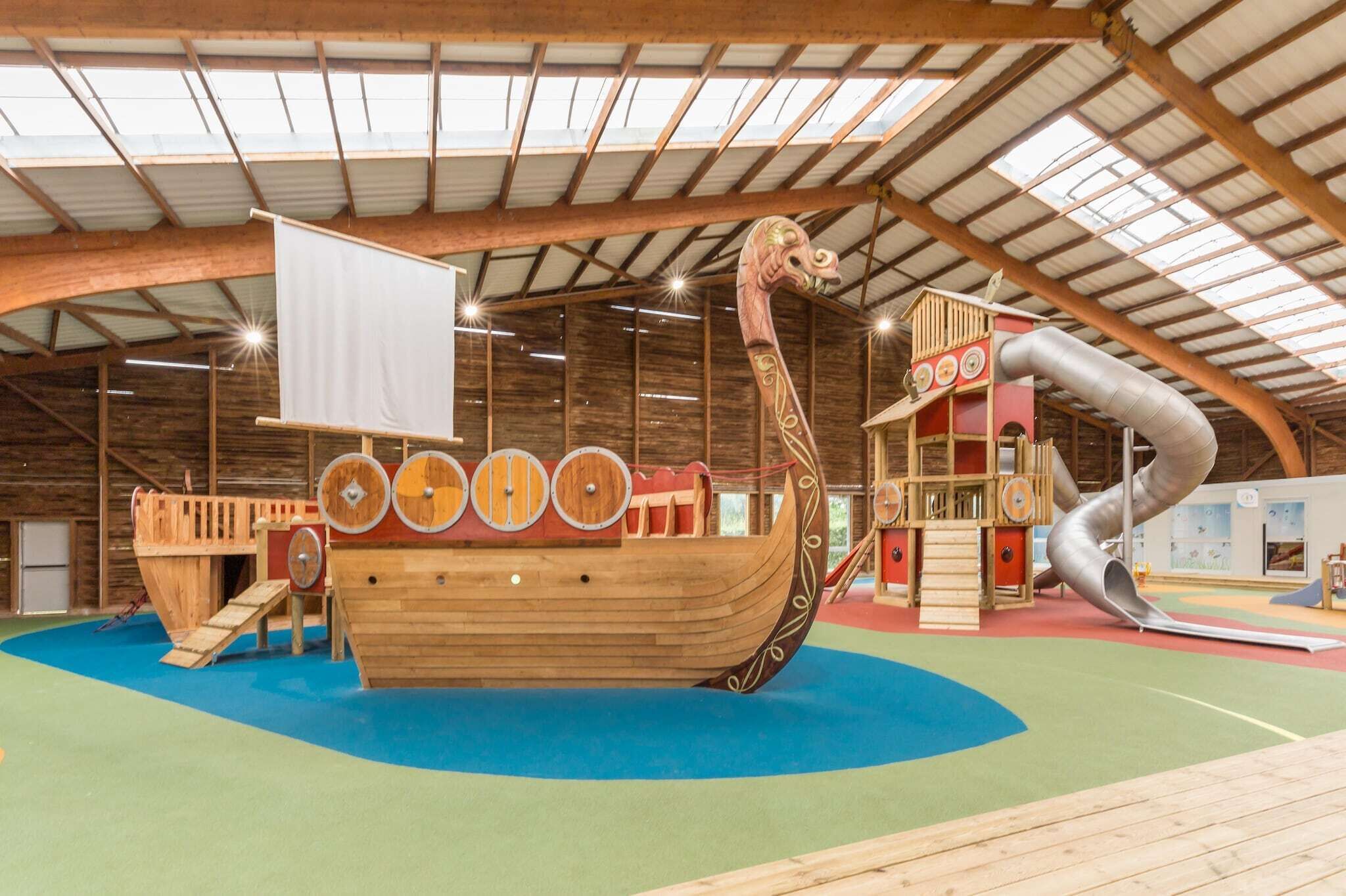 Une structure de jeu en forme de bateau en bois, dotée de toboggans et d’aires d’escalade, située à l’intérieur d’un bâtiment à plafond haut et aux poutres apparentes.