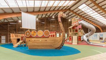 Une structure de jeu en forme de bateau en bois, dotée de toboggans et d’aires d’escalade, située à l’intérieur d’un bâtiment à plafond haut et aux poutres apparentes.