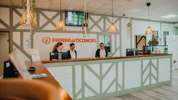 Un comptoir d’accueil avec une enseigne indiquant « Pierre & Vacances », deux employés derrière, et un téléviseur fixé au mur.