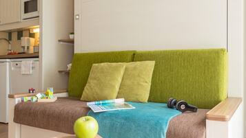 Ein kleines Zimmer mit einer Küchenzeile, einem grünen Sofa mit Kissen, einem Holztischchen mit einem Apfel und einer Zeitschrift sowie Kopfhörern auf dem Sofa.