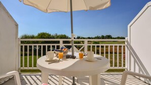 Een witte tafel met ontbijtartikelen op een balkon met een witte reling, een helderblauwe lucht en een grasveld op de achtergrond.