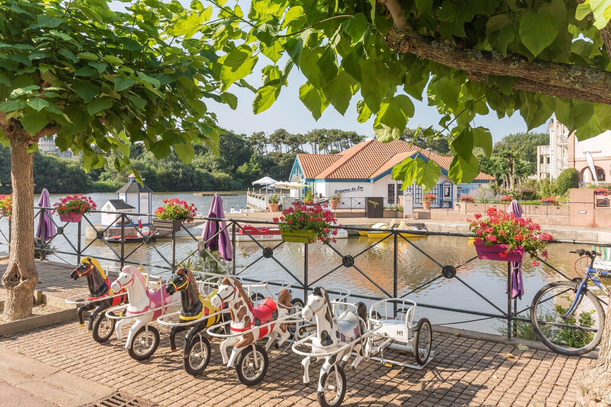 A waterfront area with a row of horse-drawn carriages, a building with a red roof, and a tree with hanging flower baskets.