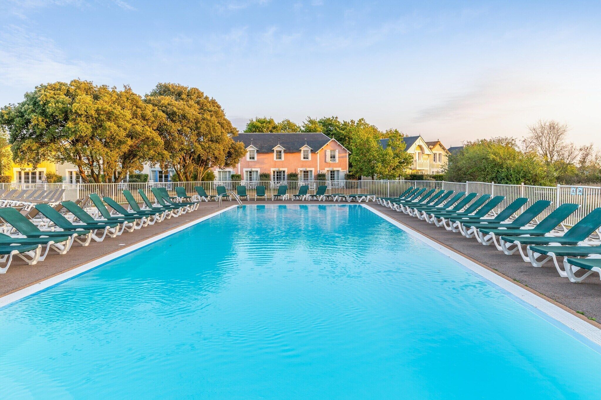 Piscina rectangular con sillas de sol verdes a ambos lados, rodeada por una valla blanca.
