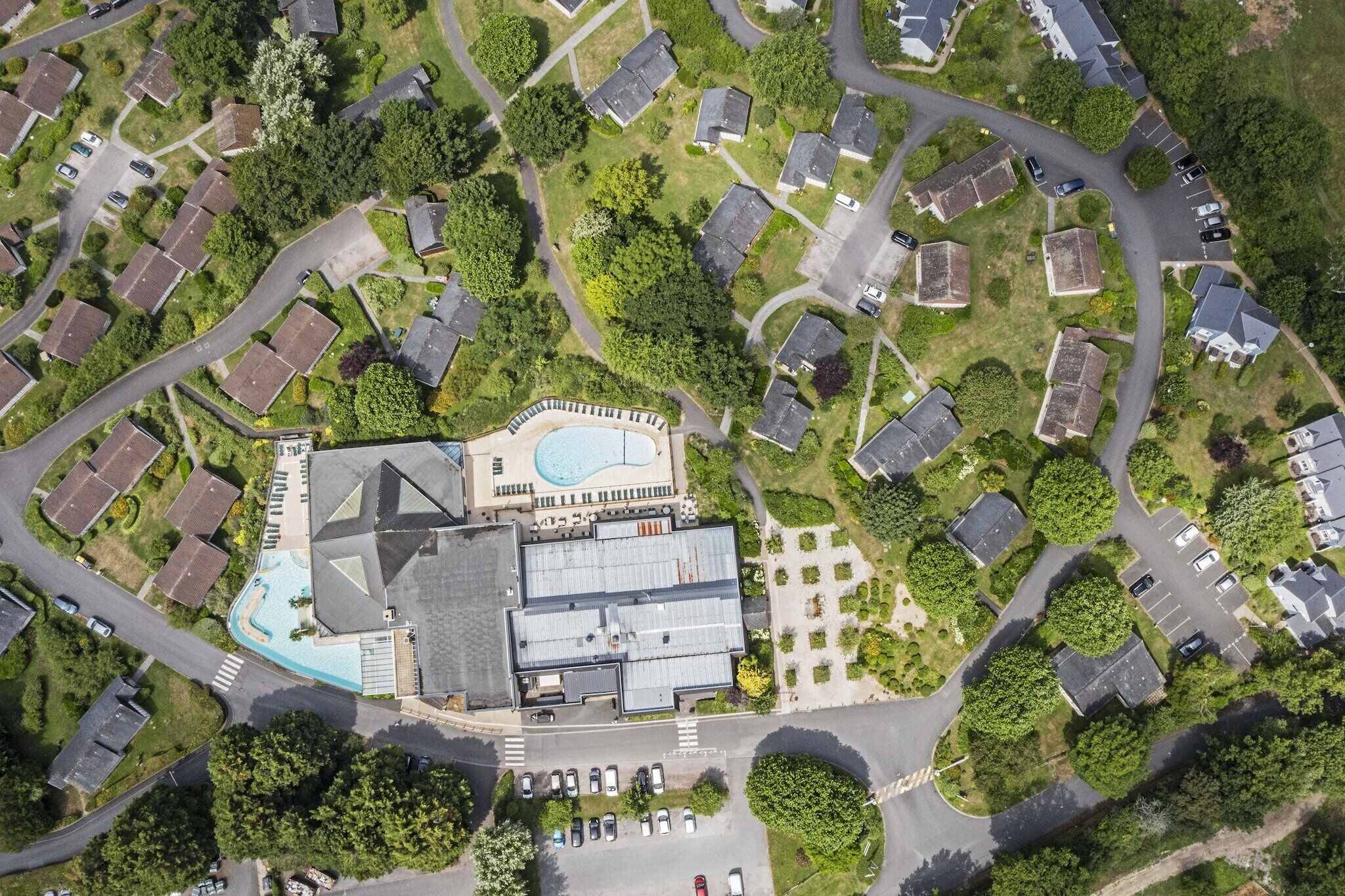 Vue aérienne d’un quartier résidentiel avec des maisons, une piscine et un grand bâtiment doté d’un toit en pente.
