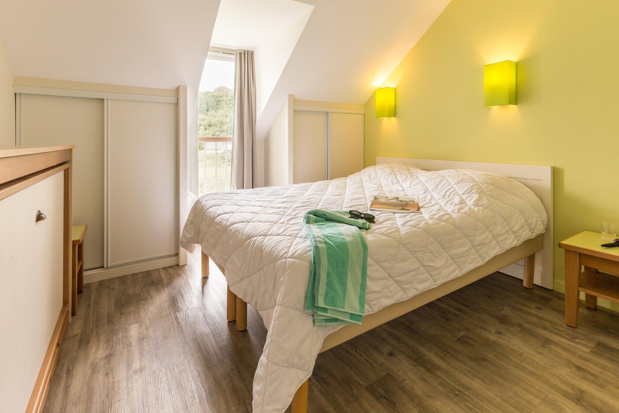 Un dormitorio con cama, mesita de noche, ventana con cortinas y lámpara de pared.