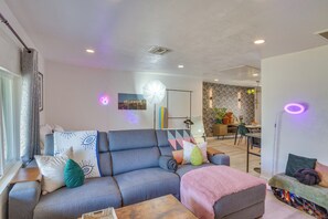 Ein modernes Wohnzimmer mit einem grauen Sofa, einem Couchtisch und einer rosafarbenen Decke. Im Hintergrund ist eine Essecke mit Tisch und Stühlen zu sehen.