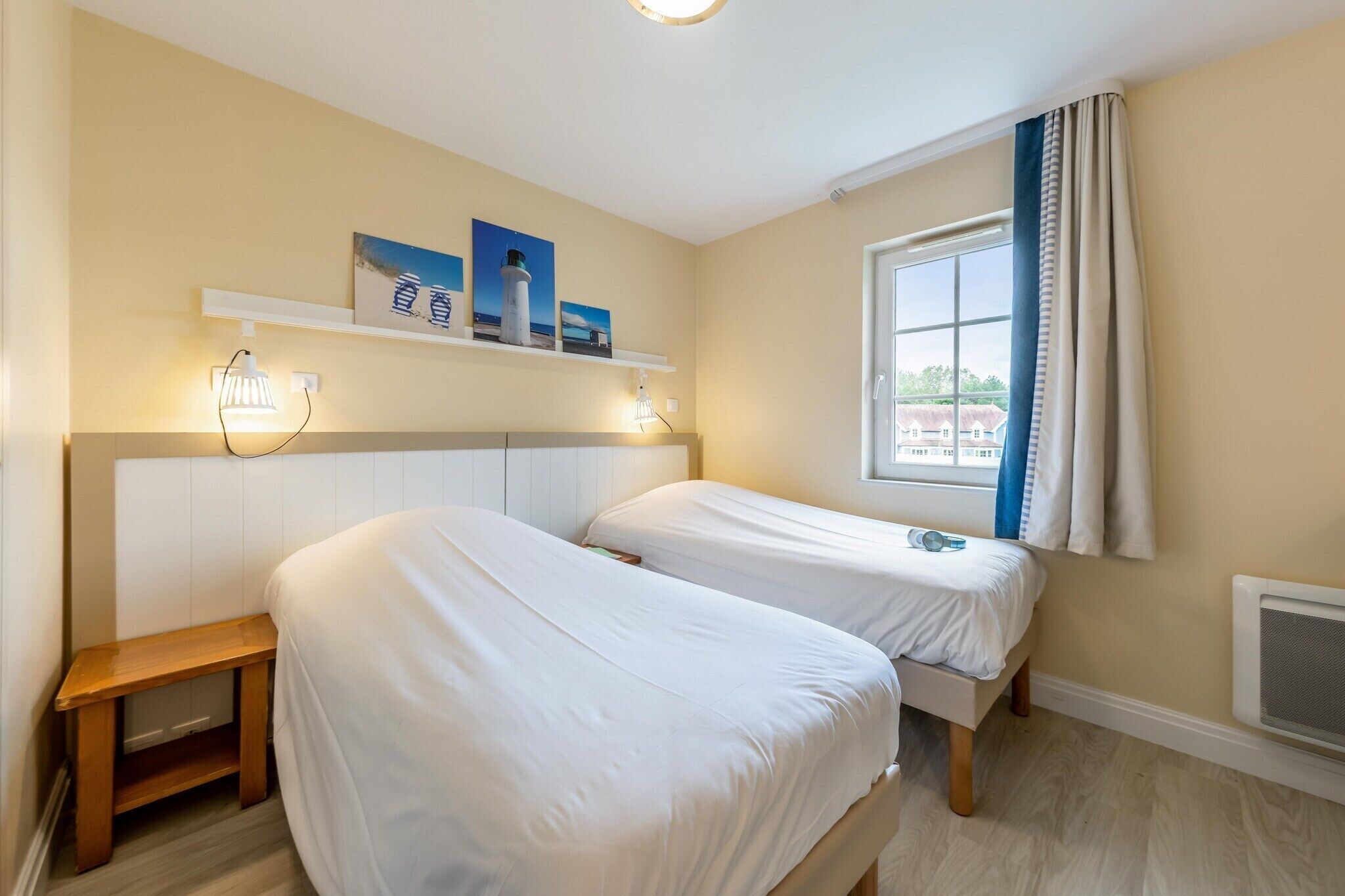 Une chambre d’hôtel avec deux lits, une petite table de chevet, une fenêtre avec des rideaux, une étagère fixée au mur avec des photos et un luminaire au plafond.