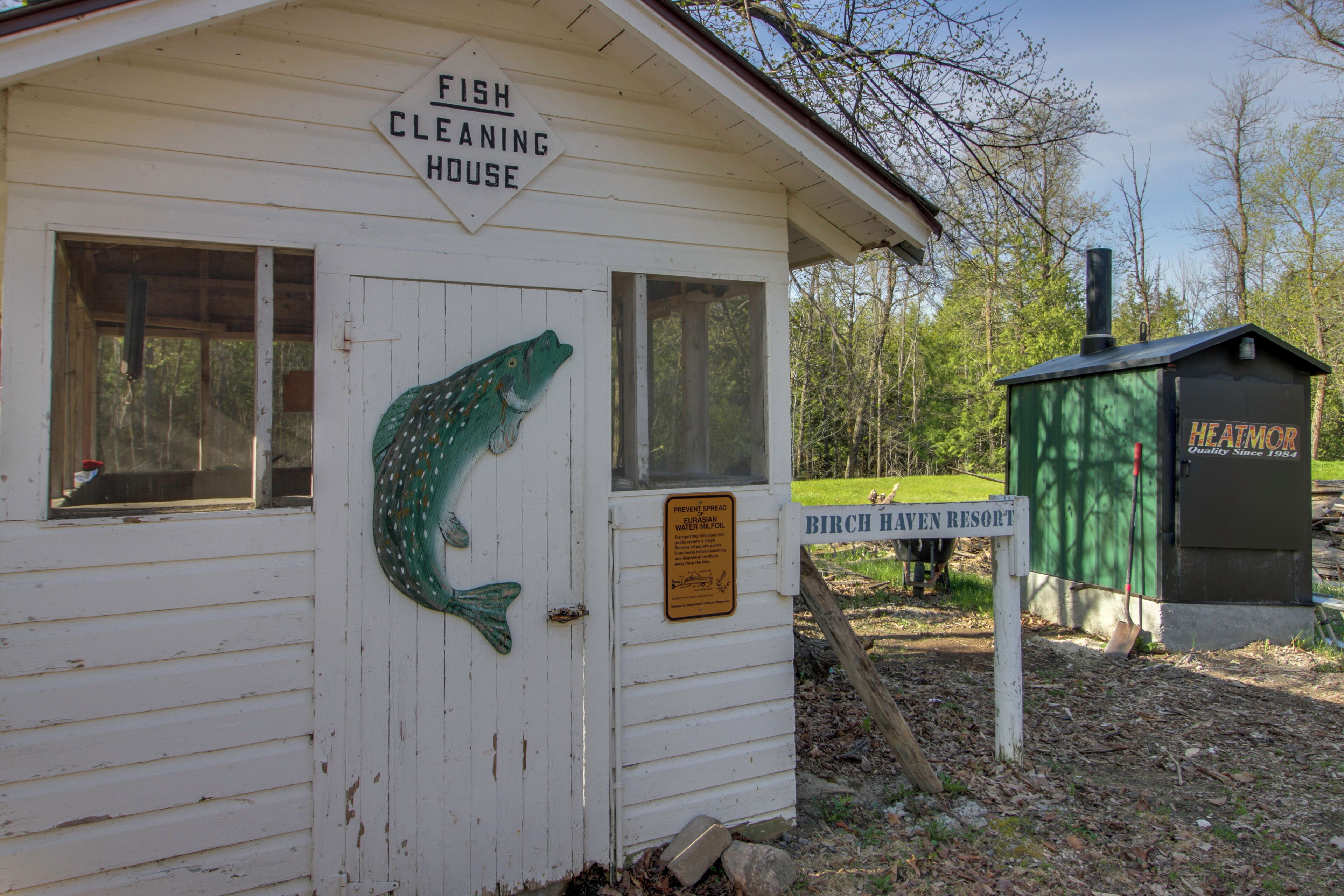 Ein weißes Gebäude mit der Aufschrift 'Fish Cleaning House', einer Fischdekoration, einem Schild für 'Birch Haven Resort' und einem grünen Verteilerkasten mit der Bezeichnung 'HEATMOR'.