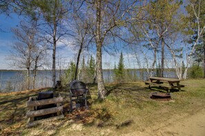 A lakeside campsite with a grill, picnic table, and fire pit.
