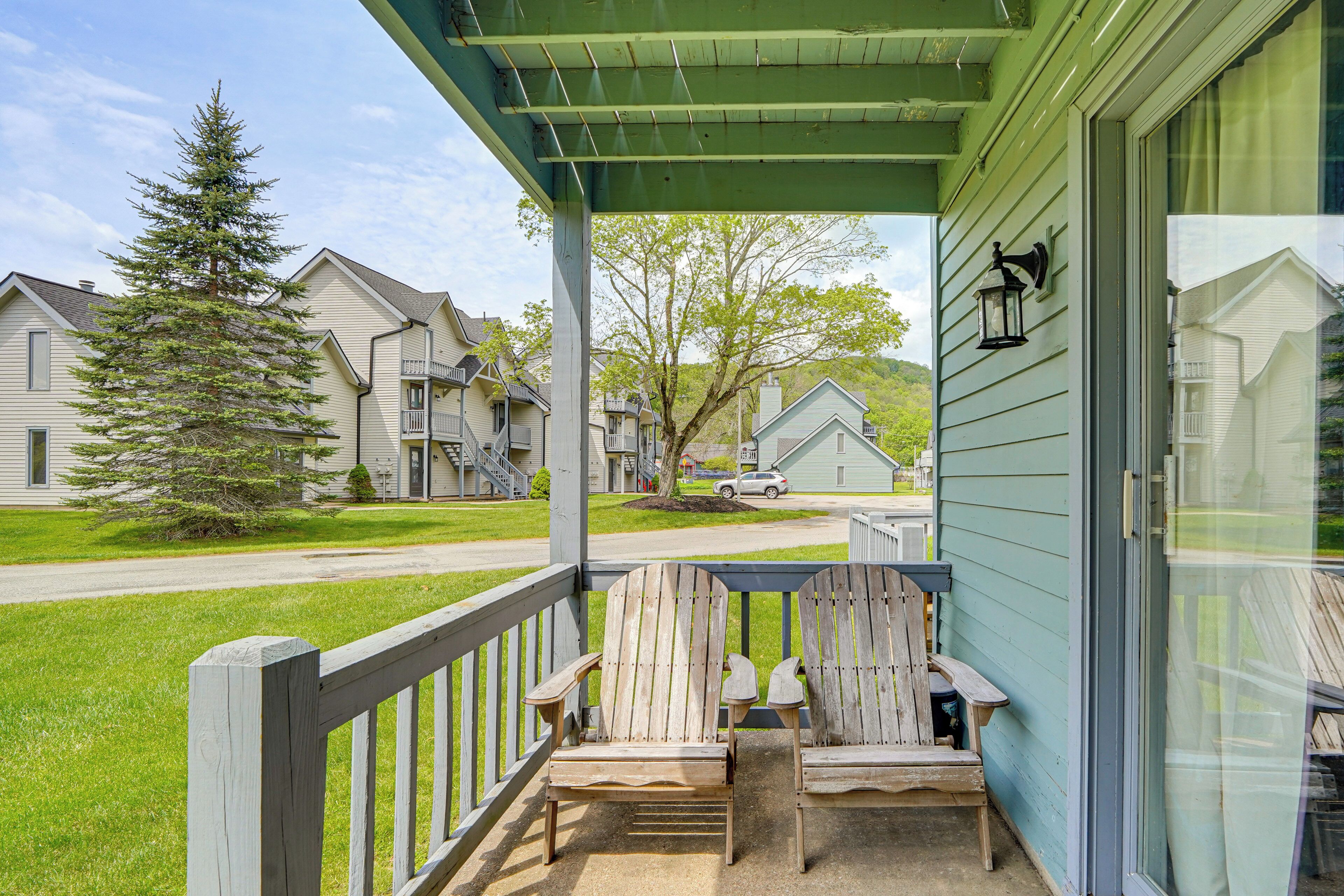 A porch with two wooden Adirondack chairs, a green awning, and a view of a residential street with houses and a tree.