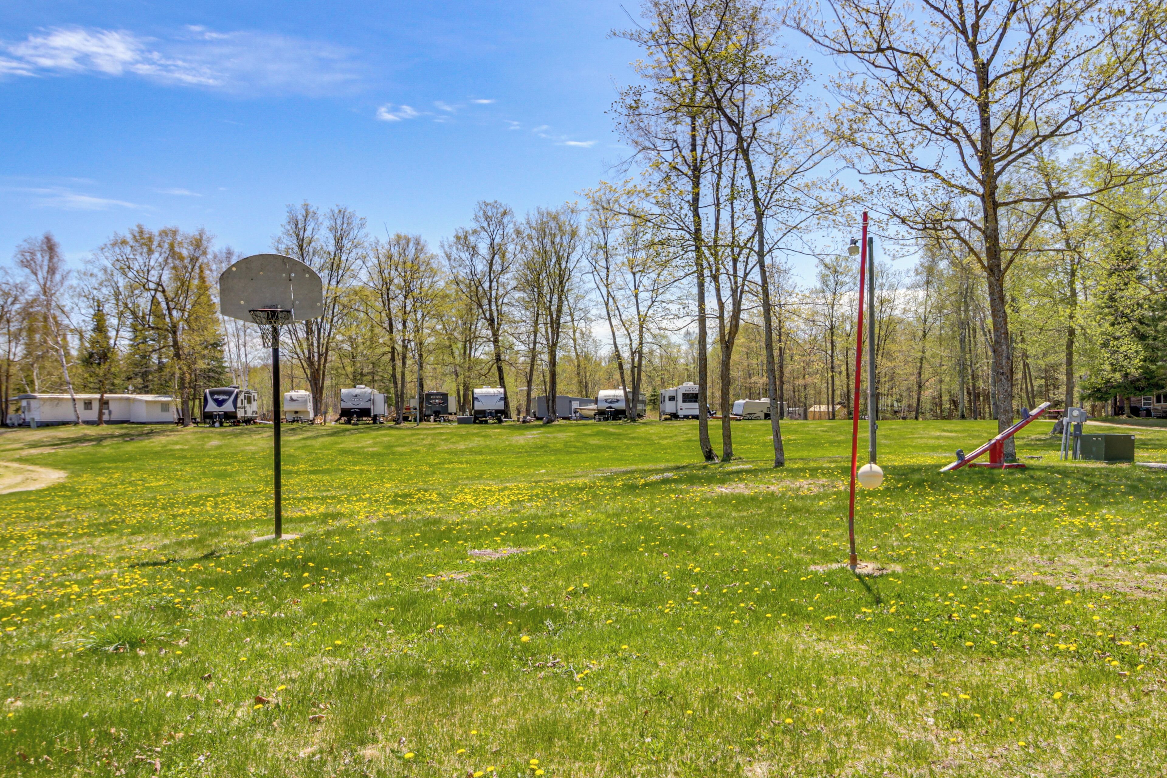 A basketball court with a hoop, trees, and RVs in the background.