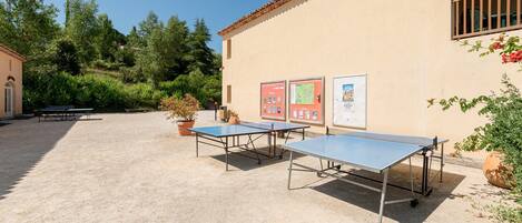 An outdoor area with ping pong tables, a building with a tiled roof, and greenery in the background.