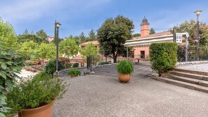 Une cour avec un chemin de gravier, des plantes en pot et un bâtiment doté d’une tour horloge.