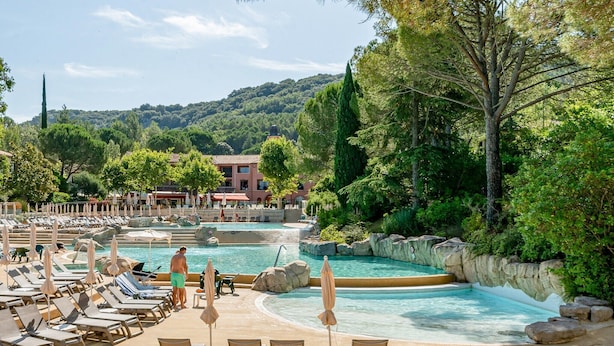 Un espace piscine doté de chaises longues et de parasols, entouré d’arbres et d’immeubles.