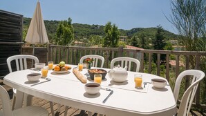 Taula preparada amb articles per esmorzar, una tetera i un bol de cafè en una terrassa de fusta amb cadires blanques.