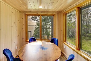 Une pièce aux murs lambrissés, avec une table ronde et des chaises bleues, de grandes fenêtres donnant sur des arbres et un bâtiment à l’extérieur.