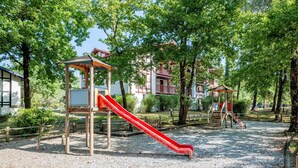 Un terrain de jeux avec des toboggans et un espace de repos couvert, entouré d’arbres et de bâtiments.
