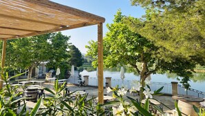 An outdoor seating area with tables and chairs, a wooden pergola, and a view of a lake. - Chic Riverfront Stay with Terrace (Mallemort)