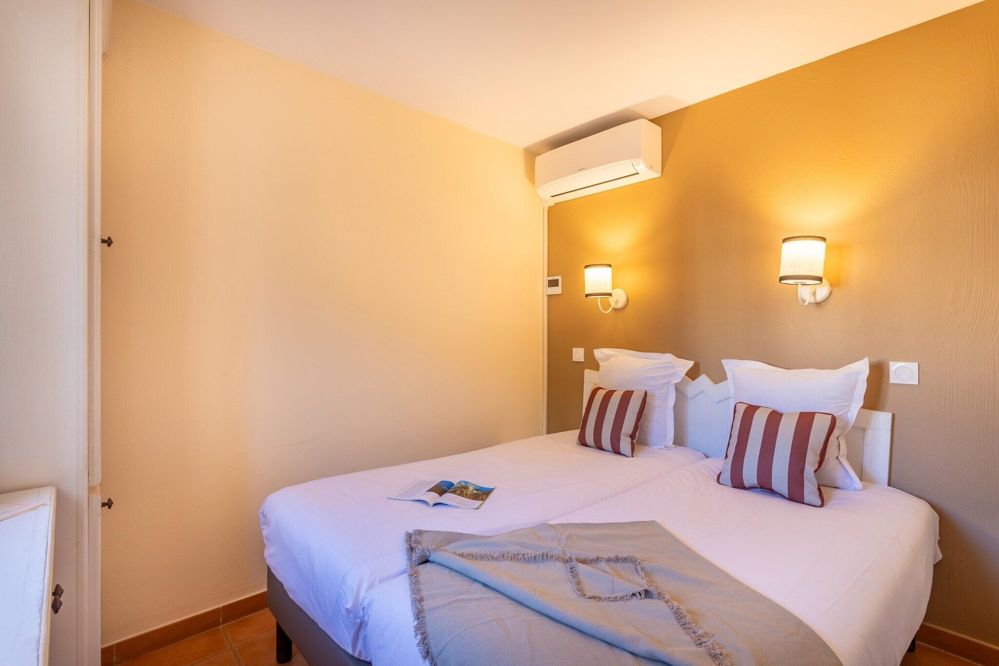 A hotel room with a bed, two wall-mounted lights, an air conditioning unit, and a door.
