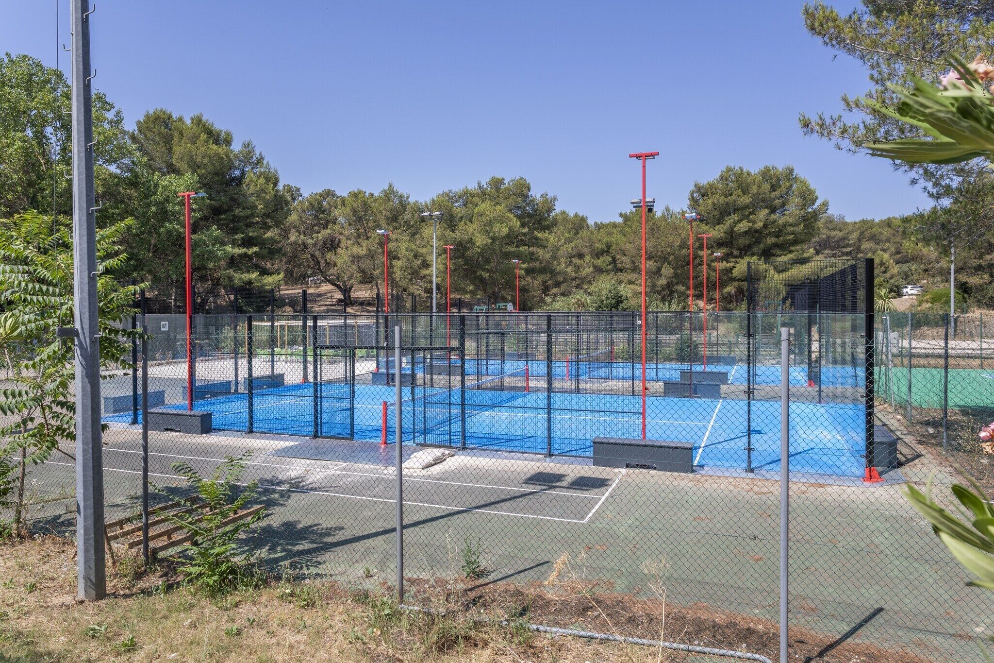 A fenced tennis court with a blue surface, surrounded by trees and a clear sky.