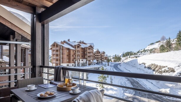 Ein Balkon mit Frühstückstisch, der Blick auf einen Skiort mit Gebäuden und verschneiter Landschaft bietet.
