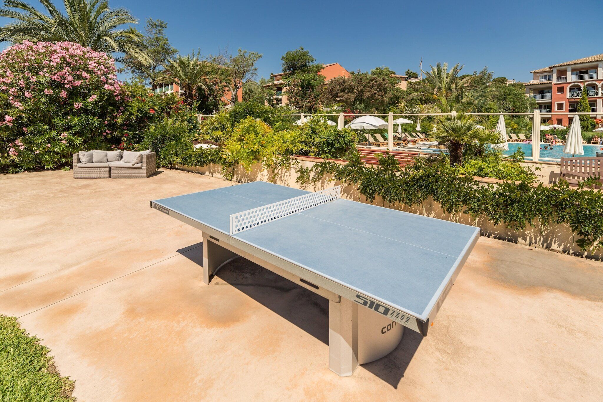 Mesa de ping pong al aire libre con red, rodeada de abundante follaje y una zona de piscina.