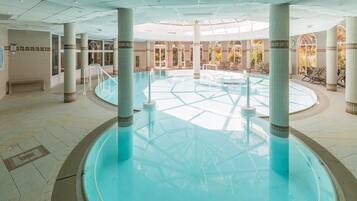 A large indoor swimming pool with a central column structure, surrounded by lounge chairs and a tiled floor.