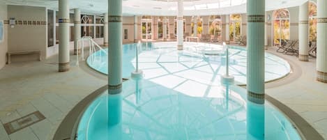 A large indoor swimming pool with a central column structure, surrounded by lounge chairs and a tiled floor.