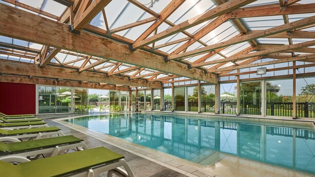 Una piscina interior con vigas de madera y paredes de vidrio, rodeada de sillones de descanso.