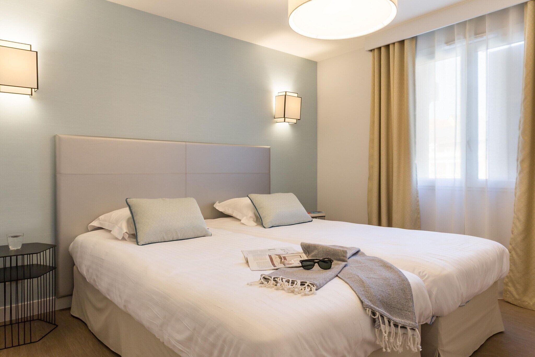 Une chambre d’hôtel avec un grand lit, deux tables de chevet, une lampe fixée au mur et une fenêtre avec des rideaux.