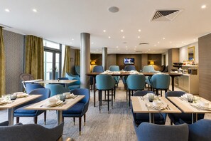 Un comedor de hotel con mesas dispuestas para comidas, sillas azules, un televisor y una encimera de cocina.