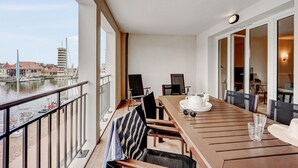 Un balcone con tavolo e sedie di legno, vista sull'acqua e sugli edifici, e una porta vetrata che dà su un ambiente interno.