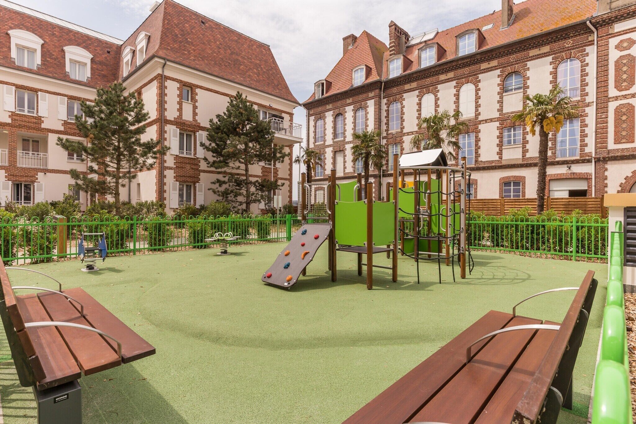 Une aire de jeux avec des structures d’escalade et un toboggan, entourée de bancs et d’une clôture verte.