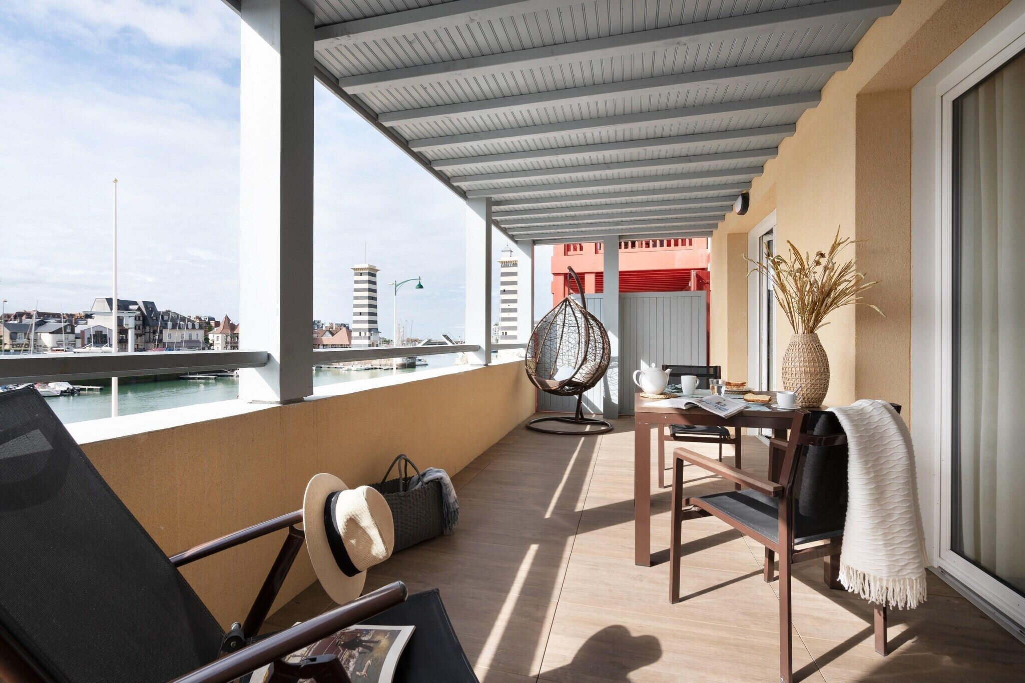 A balcony with a view of the marina, a hanging chair, a table with a vase, and a chair with a throw blanket.