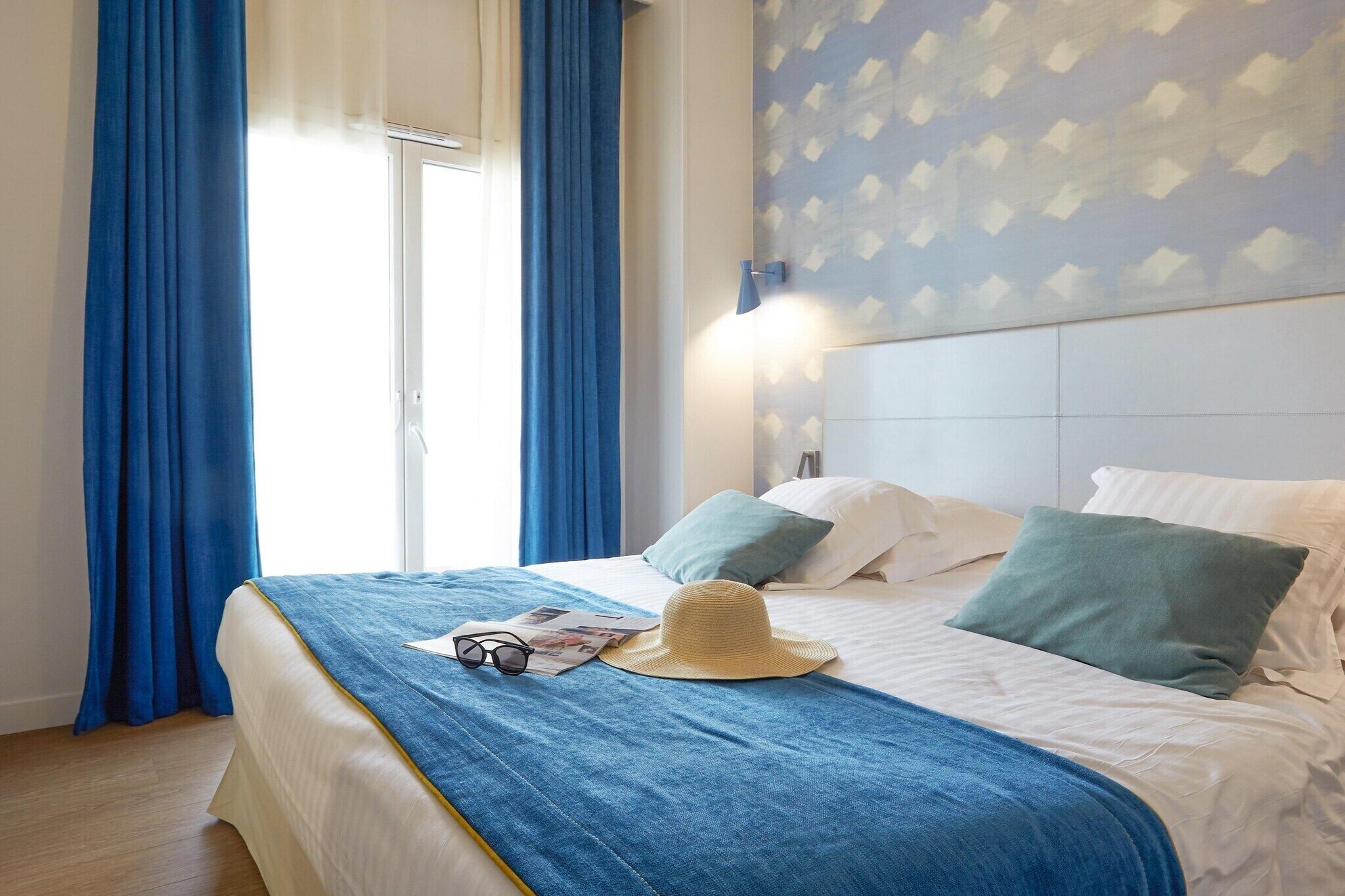 Een hotelkamer met een bed, blauwe gordijnen, een hoed en een tijdschrift.