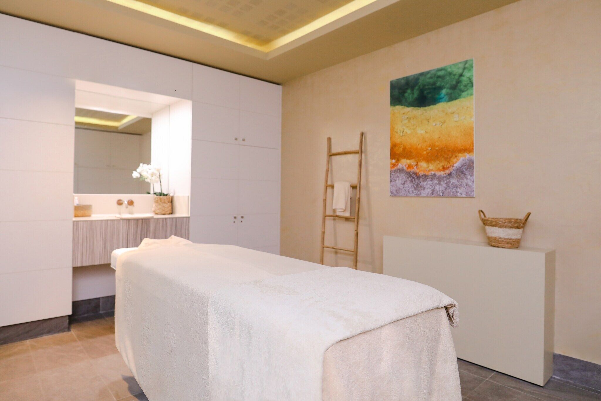 A spa room with a massage table, a large mirror, a painting, and a small shelf with a basket.