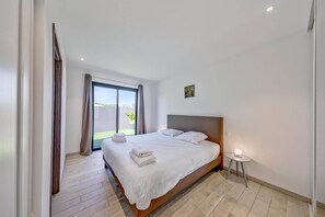 A modern hotel room with a large bed, a small bedside table with a lamp, a window with curtains, and a view of the outdoors. - Lecci Retreat near Cala Rossa Beach (Porto-Vecchio)