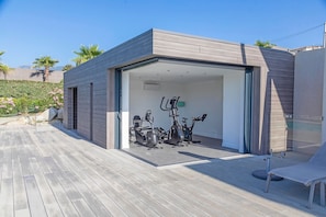 Ein modernes Fitnessstudio im Freien mit Trainingsgeräten, einer Holzterrasse und einer Glastür zu einem überdachten Bereich.