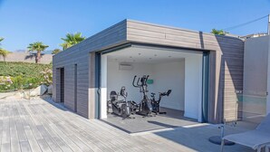 Ein modernes Fitnessstudio im Freien mit Trainingsgeräten, einer Holzterrasse und einer Glastür zu einem überdachten Bereich.
