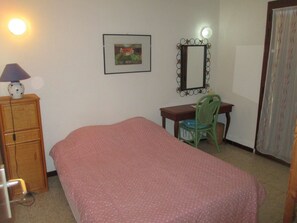 Ein Schlafzimmer mit Bett, Schreibtisch, Stuhl, Kommode, Lampe, einem gerahmten Bild und einem Spiegel.