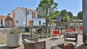 Een wit gebouw met oranje luiken, een omheind terras met rieten meubels, en een grote potplant.