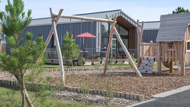 Een speeltuin met een schommel en klimconstructies, omgeven door een aangelegde tuin met houtsnippers en jonge bomen.