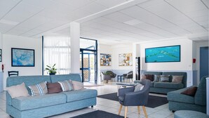 Ein geräumiger Wohnbereich mit einem blauen Sofa, einem grauen Sessel und einer blauen Couch. Ein großes Fernsehgerät an der Wand zeigt eine Karte.